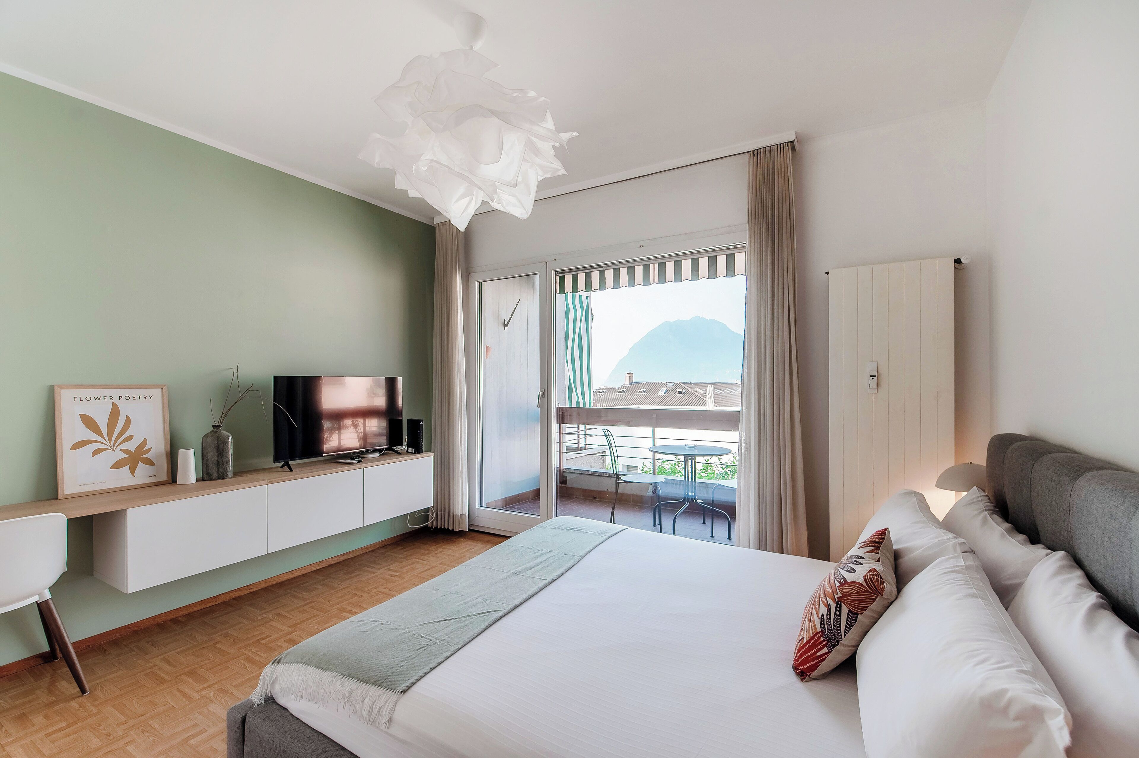 A bedroom with a bed, a TV on a stand, a desk, and a balcony with a view of the mountains.