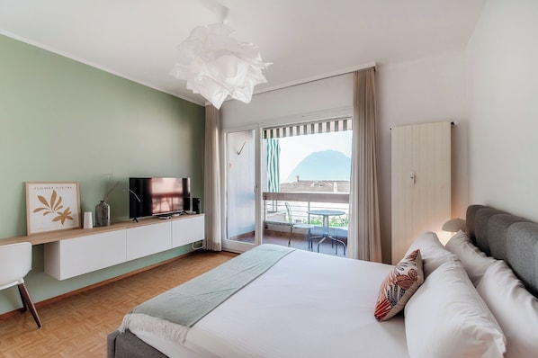 A bedroom with a bed, a TV on a stand, a desk, and a balcony with a view of the mountains. - Vinorum Studio (Massagno)
