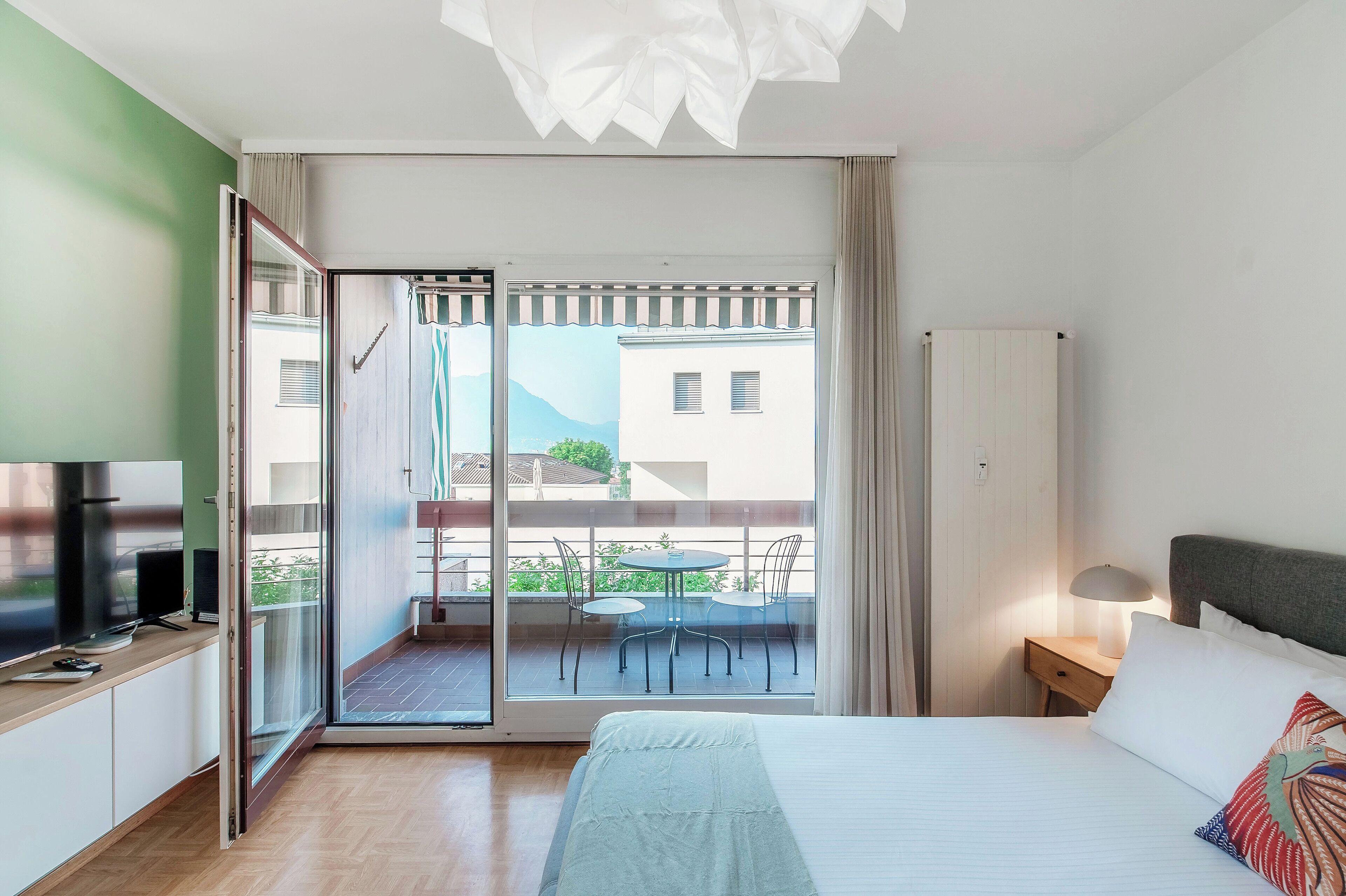 A modern hotel room with a bed, bedside table, lamp, and a balcony with a table and chairs.