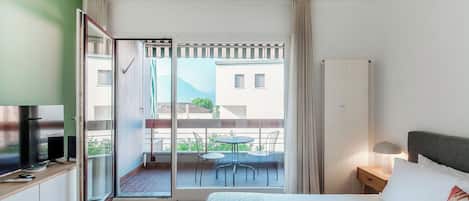 A modern hotel room with a bed, bedside table, lamp, and a balcony with a table and chairs.