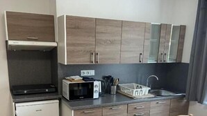 Una cocina compacta con microondas, tostadora y lavabo. Hay un refrigerador pequeño y armarios de madera. Un comedor con sillas se ve en primer plano.