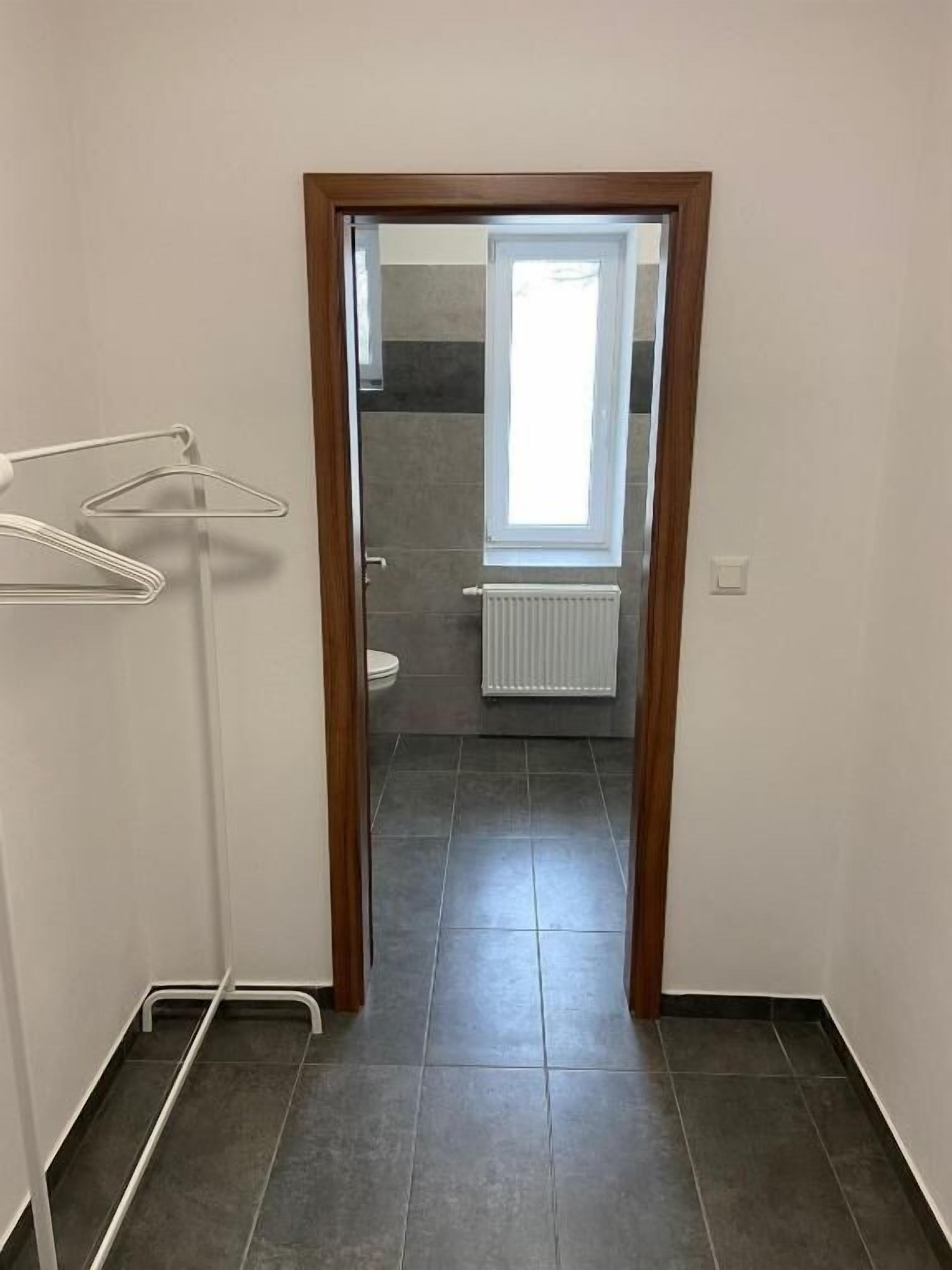 Un couloir étroit avec une porte en bois donnant sur une salle de bain équipée d’une fenêtre et d’un radiateur.