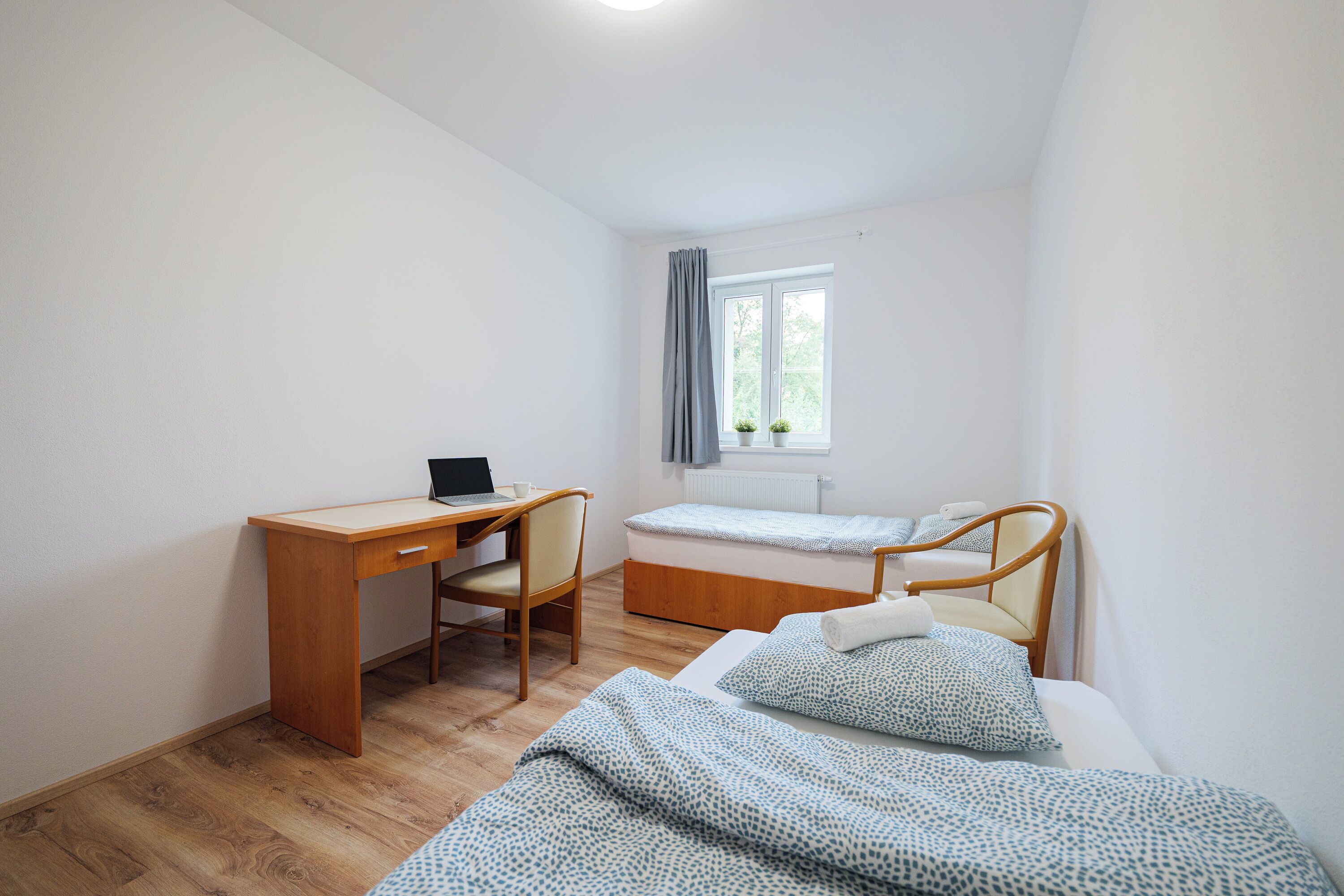 Quarto com escrivaninha de madeira, um laptop, cama com roupa de cama estampada e janela com cortinas.
