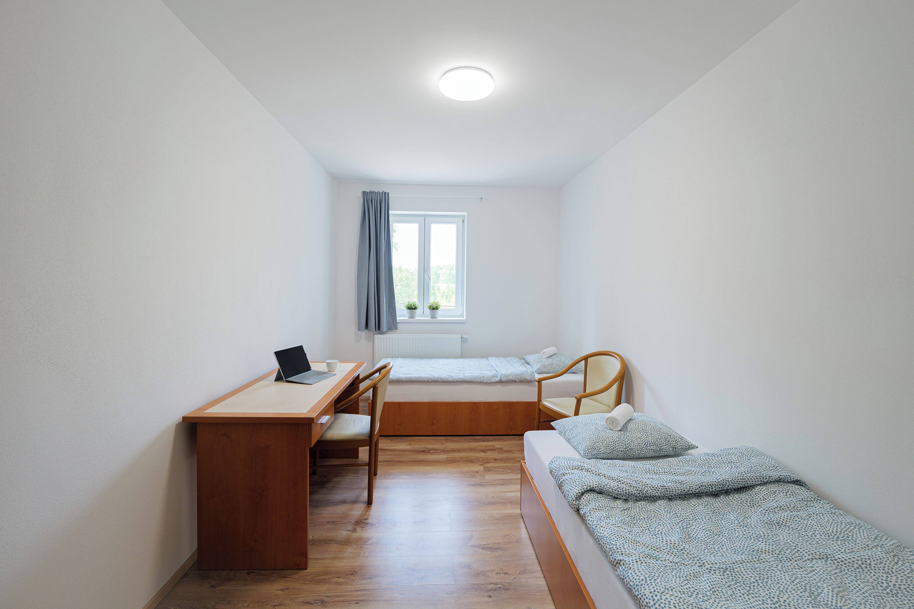 Una habitación pequeña y bien iluminada con un escritorio, una silla y dos camas individuales.