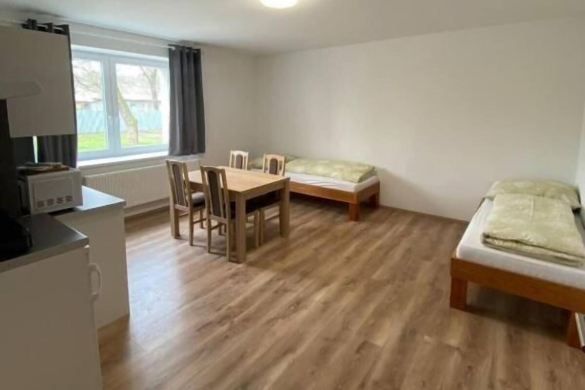 A compact room with a wooden floor, a small dining table with chairs, a single bed, and a kitchenette area with a microwave.