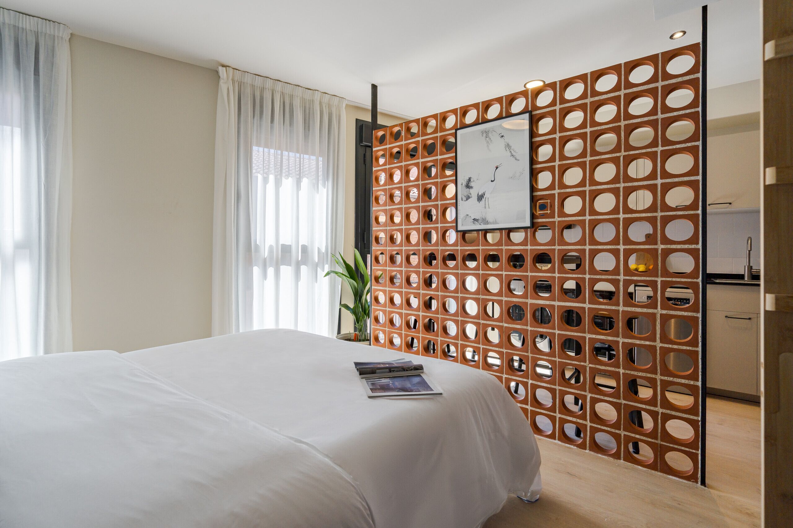 A hotel room with a large bed, a wooden partition with circular cutouts, a framed artwork, and a plant.