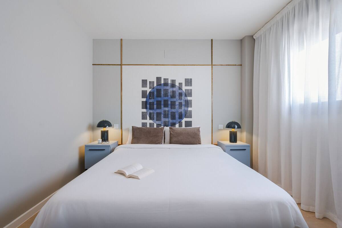 Un lit bien fait, avec un livre posé dessus, une œuvre d’art moderne et abstraite accrochée au mur, et deux tables de chevet avec des lampes.