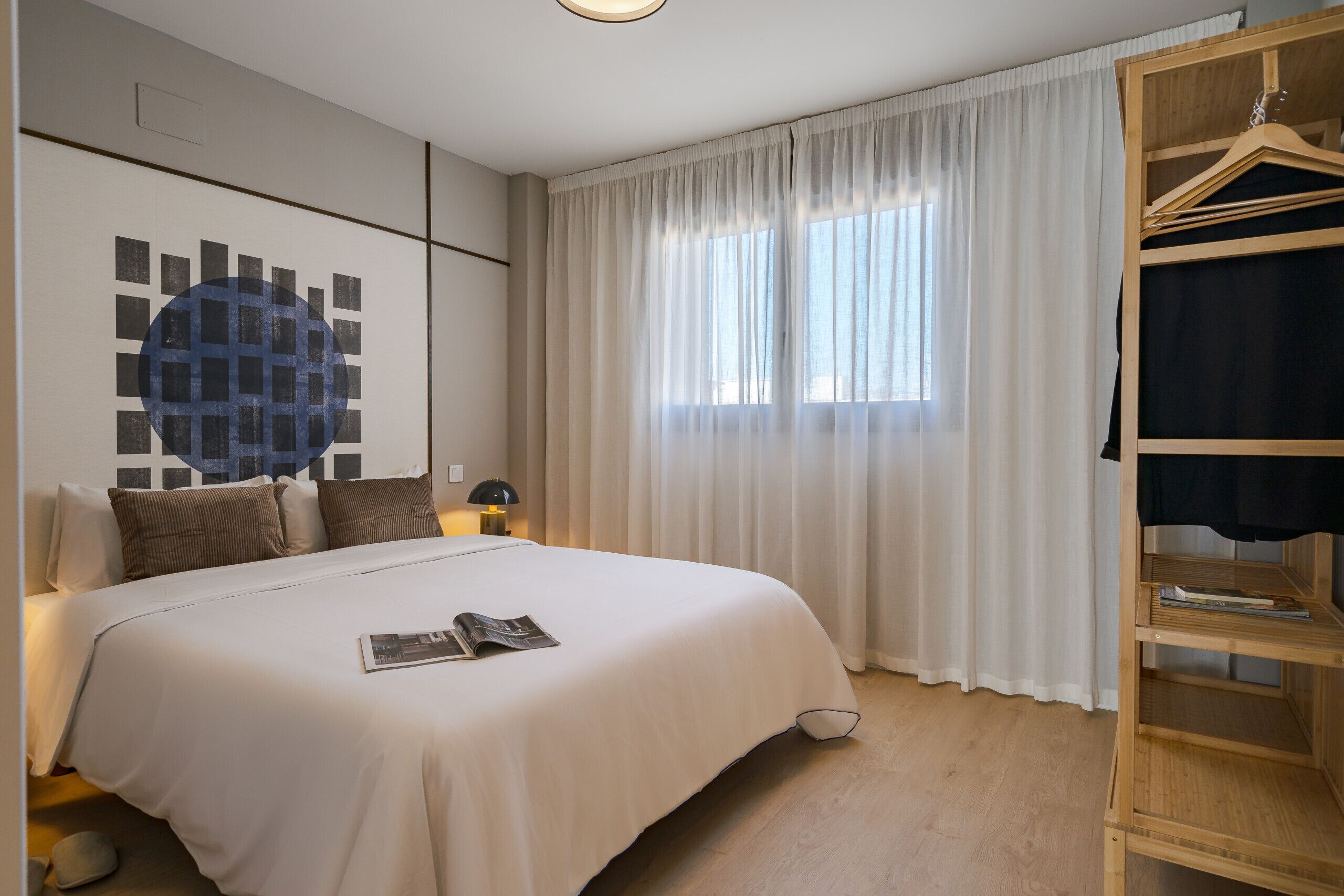 Een verzorgde slaapkamer met een groot bed, een nachtkastje, een leeslamp, een kledingkast en een raam met dunne gordijnen.