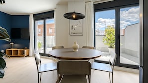 Un espace de repas moderne avec une table ronde, six chaises, un luminaire suspendu et une vue sur la ville grâce à de grandes fenêtres.