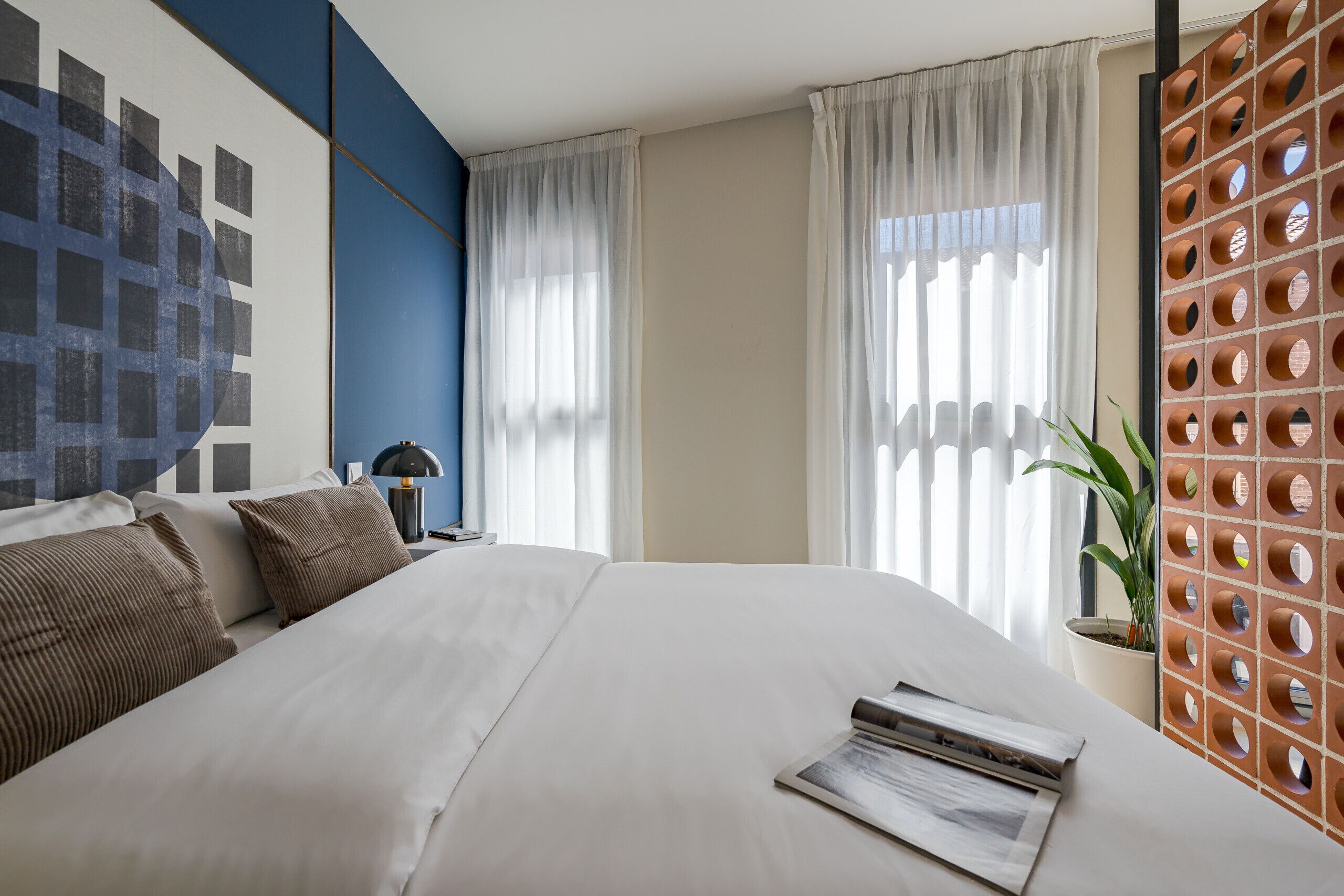 A modern hotel room with a large bed, a wall art piece, a potted plant, and a unique brick-patterned partition.
