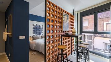 Una habitación moderna con pared azul de acento, un separador de madera con diseños circulares, una cama, una mesita con taburetes y un ventanal con vista a edificios.