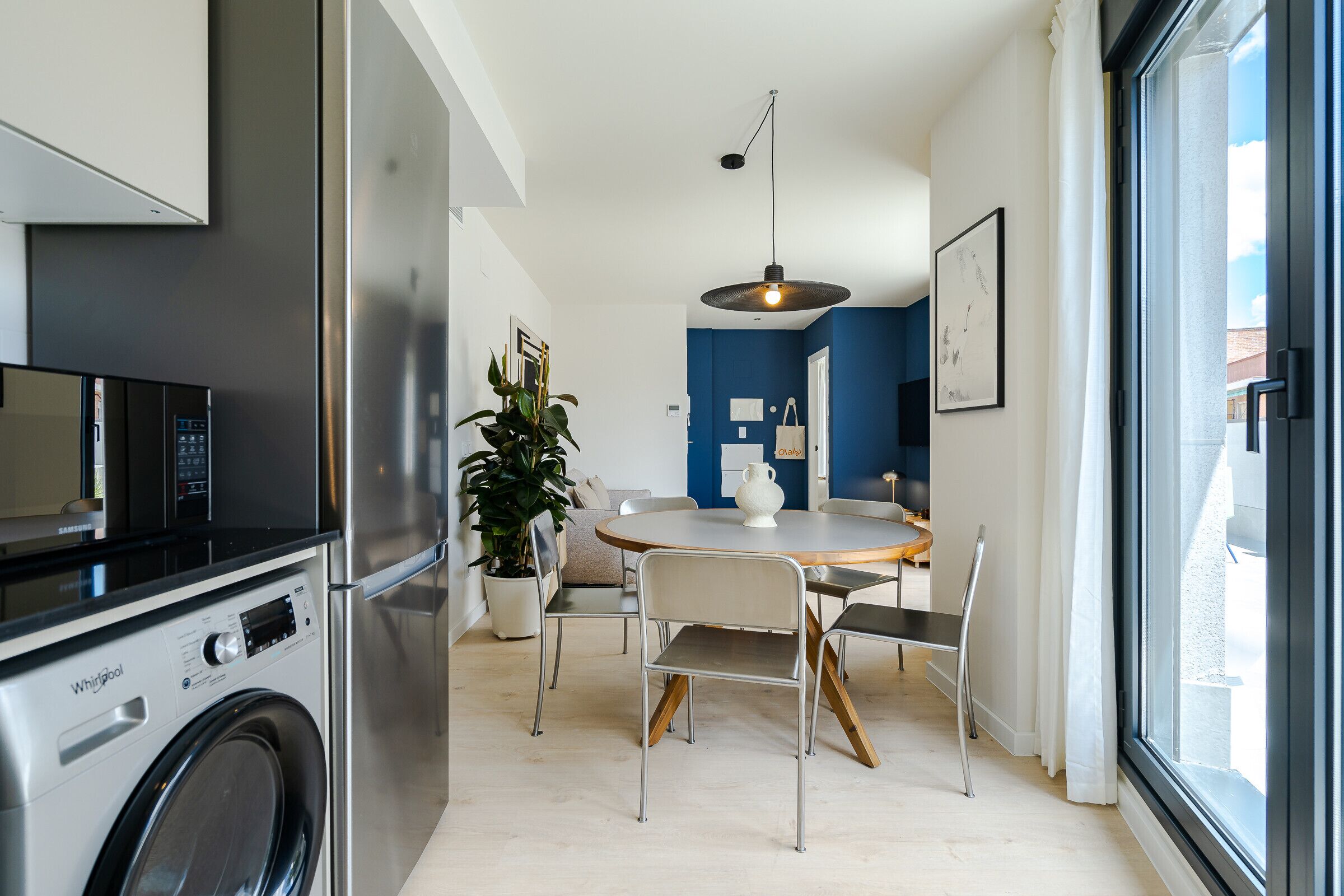 A modern kitchen with a built-in washer and dryer, a dining area with a round table and chairs, and a large window offering a view of the outside.
