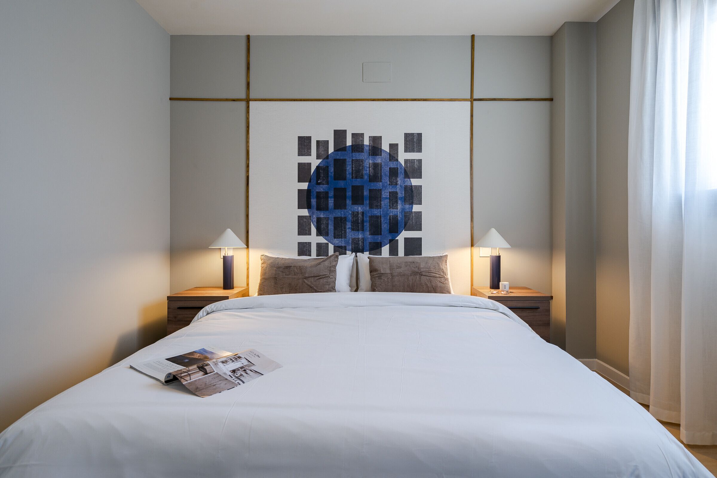 A neatly made bed with a magazine on it, two bedside lamps, and a wall art piece above the bed.