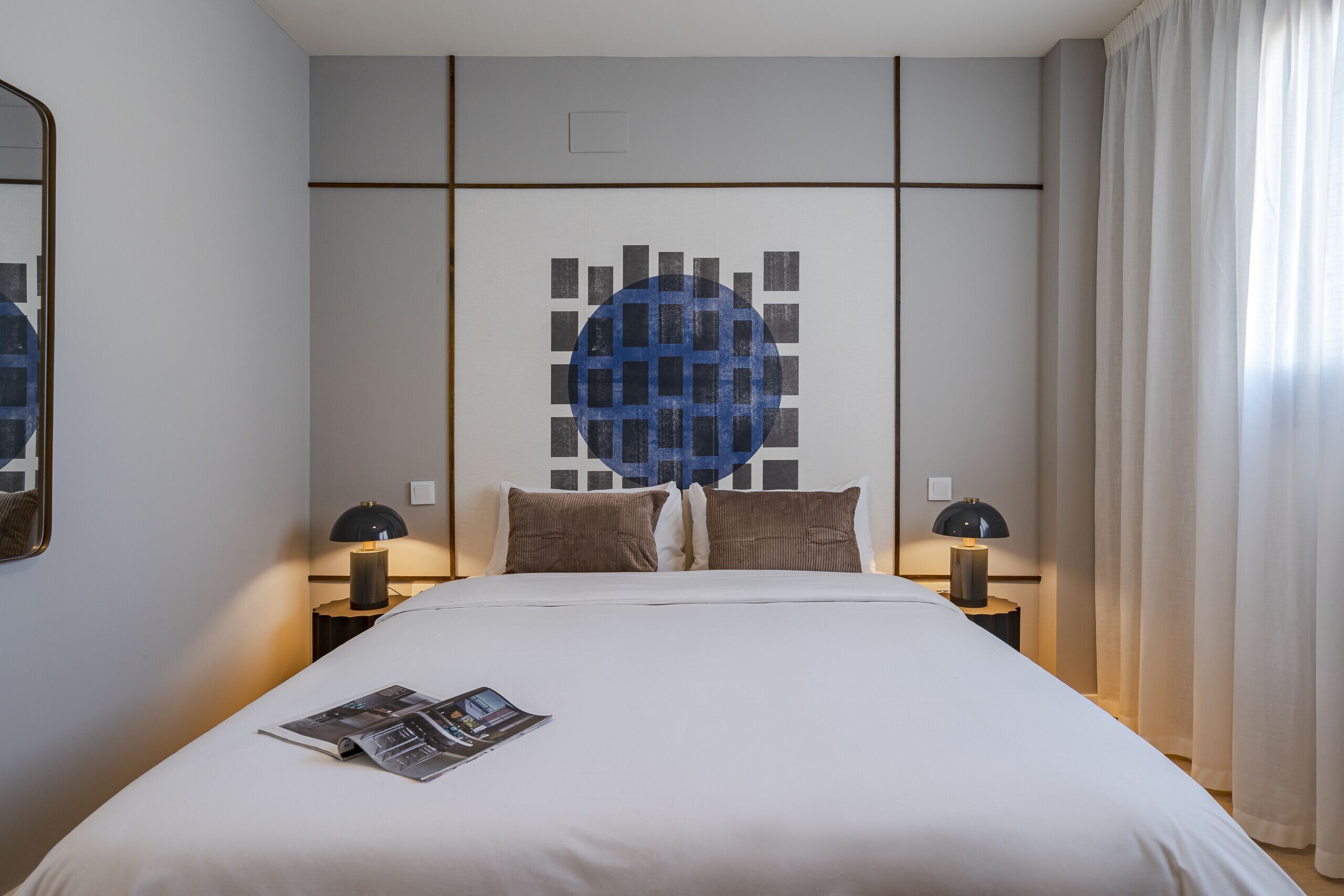 Una cama bien hecha con una revista, dos lámparas de noche y una obra de arte en la pared.