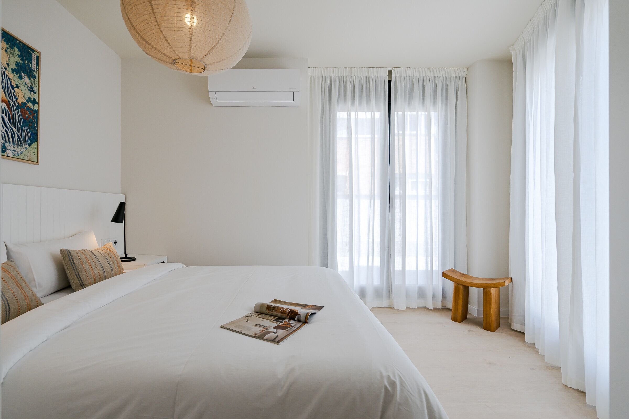 Un dormitorio con una cama grande, una mesita de noche con lámpara, un banco de madera y un ventanal con cortinas ligeras.