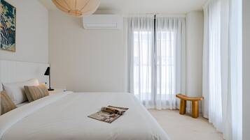 Un dormitorio con una cama grande, una mesita de noche con lámpara, un banco de madera y un ventanal con cortinas ligeras.