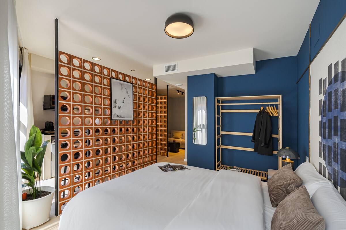 Une chambre d’hôtel moderne avec un grand lit, une cloison en bois ornée d’ouvertures circulaires, une armoire et une plante en pot.