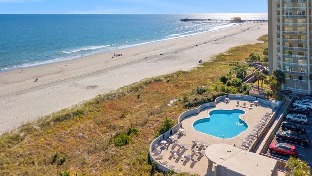 Une vue sur la plage avec une piscine et des chaises longues, une jetée en arrière-plan et un bâtiment d’hôtel.