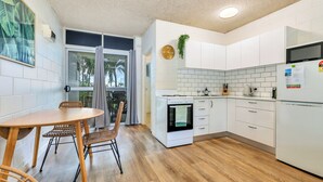 A modern kitchen with white cabinets, a wooden dining table with chairs, and a view through a glass door. - Self contained studio apartment overlooking the beautiful Nightcliff foreshore with your own private balcony. (Nightcliff)