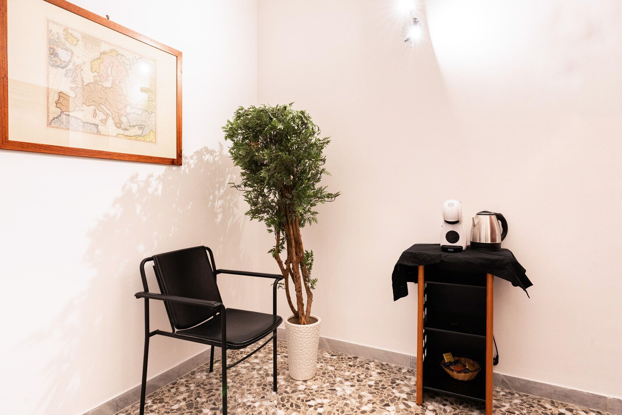 Une pièce avec une chaise noire, une plante en pot, une petite table avec une bouilloire et une carte encadrée accrochée au mur.