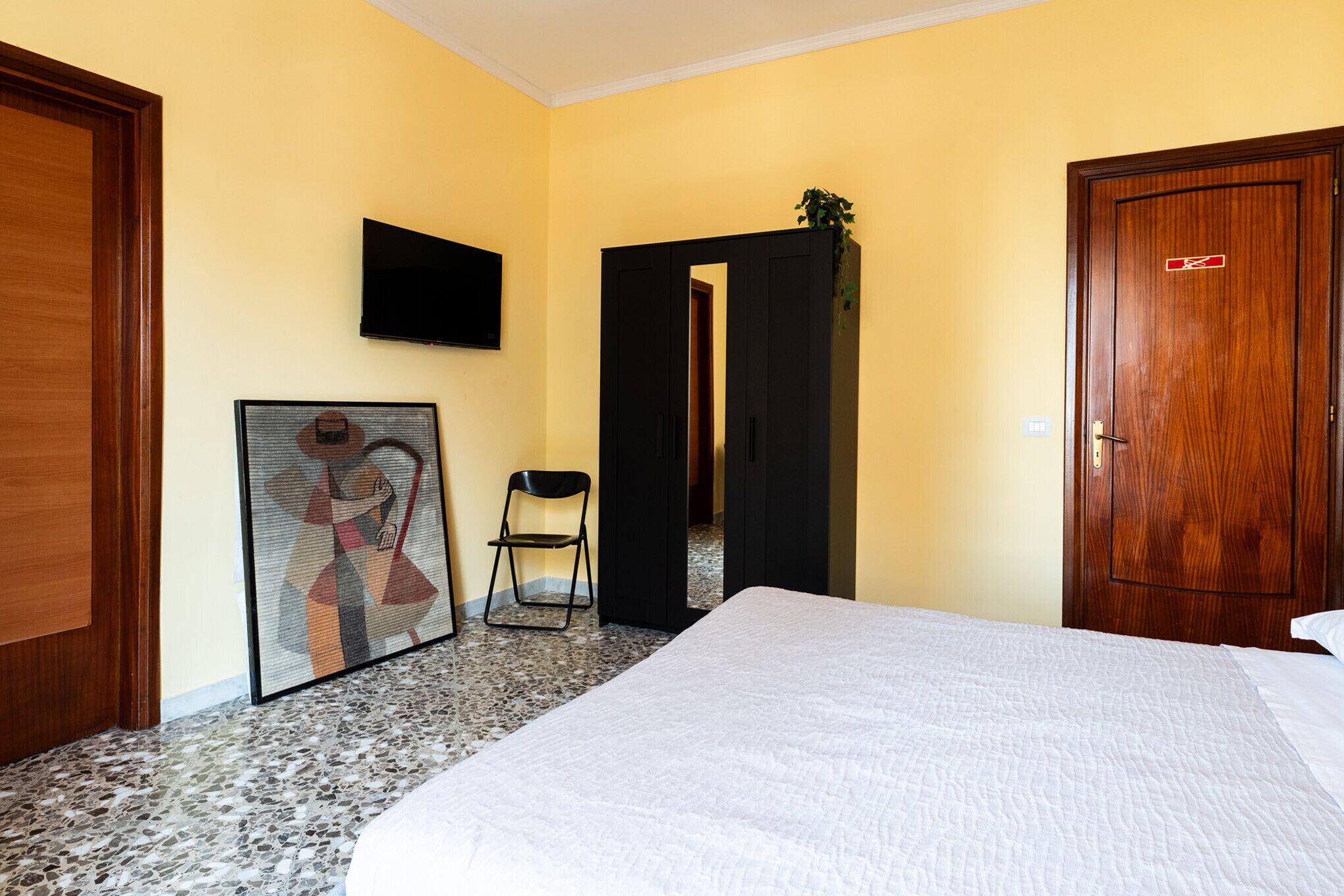 Habitación de hotel con cama, televisión montada en la pared, un cuadro enmarcado, una silla y una puerta.