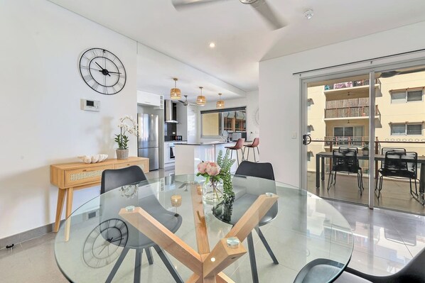 A modern kitchen with stainless steel appliances, a dining area with a glass table and chairs, and a view of the outside balcony. - Darwin Inner City Oasis | 2 Bed Apt (Darwin City)
