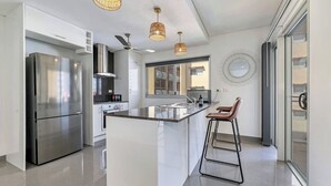 A modern kitchen with a stainless steel refrigerator, a central island with bar stools, pendant lighting, and a large window providing natural light. - Darwin Inner City Oasis | 2 Bed Apt (Darwin City)