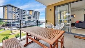A balcony with a wooden table and chairs, a glass door leading to a living area, and a view of modern buildings across a body of water. - Waterfront Pearl Suite: 2 Bed Luxury Apt by Tropical BNB (Darwin City)