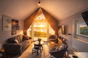 Quarto acolhedor com teto inclinado, um sofá, uma poltrona, uma mesa pequena e vista para o exterior.