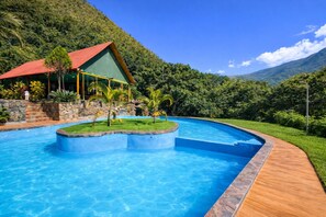 Una piscina con cubierta de madera, un edificio con techo rojo y abundante vegetación al fondo.