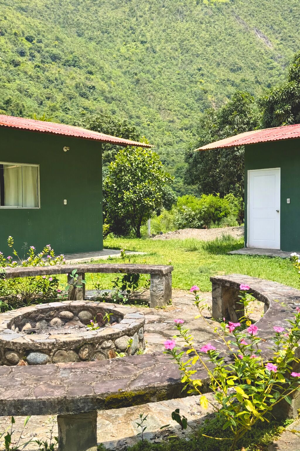 Dos edificios verdes con techos rojos, un hogar de piedra y flores rosadas en un jardín.