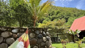A hammock tied between rocks with a stone wall, a palm tree, and a house with a red roof in the background. - www.ecoterralodge.com (Santa Teresa)
