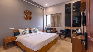 A modern bedroom with a wooden bed, a small side table, a desk with a chair, a ceiling fan, and a large window with curtains. - Kailasham Stay (Dharamshala)