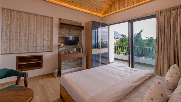A bedroom with a large bed, a TV on a wooden shelf, a balcony with a view of trees and mountains, and a wooden ceiling. - Kailasham Stay (Dharamshala)