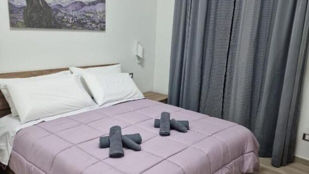 A neatly made bed with a purple bedspread and rolled towels, a white wall with a framed artwork, and grey curtains. - Greta family house: Clare (Roma)