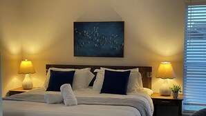 Una cama bien tendida con ropa de cama blanca y azul, dos lámparas de noche, un cuadro colgado en la pared y una ventana con persianas.