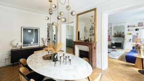A dining area with a marble table, black chairs, and a chandelier. There is a fireplace and a bookshelf in the background. - ELEGANT APARTMENT IN THE HEART OF PARIS – STEPS FROM JARDIN DU LUXEMBOURG (Paris)