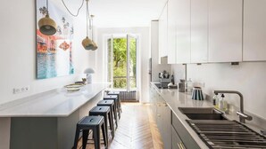 A modern kitchen with a large island, bar stools, a sink, and a view of greenery outside. - ELEGANT APARTMENT IN THE HEART OF PARIS – STEPS FROM JARDIN DU LUXEMBOURG (Paris)