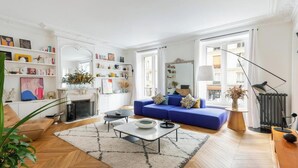 A modern living room with a blue sofa, a fireplace, and a bookshelf filled with books and decorative items. - ELEGANT APARTMENT IN THE HEART OF PARIS – STEPS FROM JARDIN DU LUXEMBOURG (Paris)