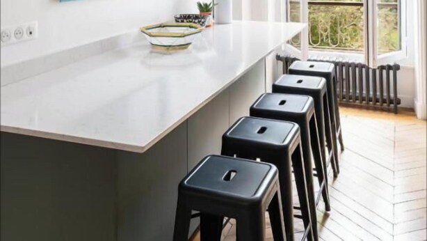 A modern kitchen with a white countertop, black bar stools, and pendant lights. - ELEGANT APARTMENT IN THE HEART OF PARIS – STEPS FROM JARDIN DU LUXEMBOURG (Paris)