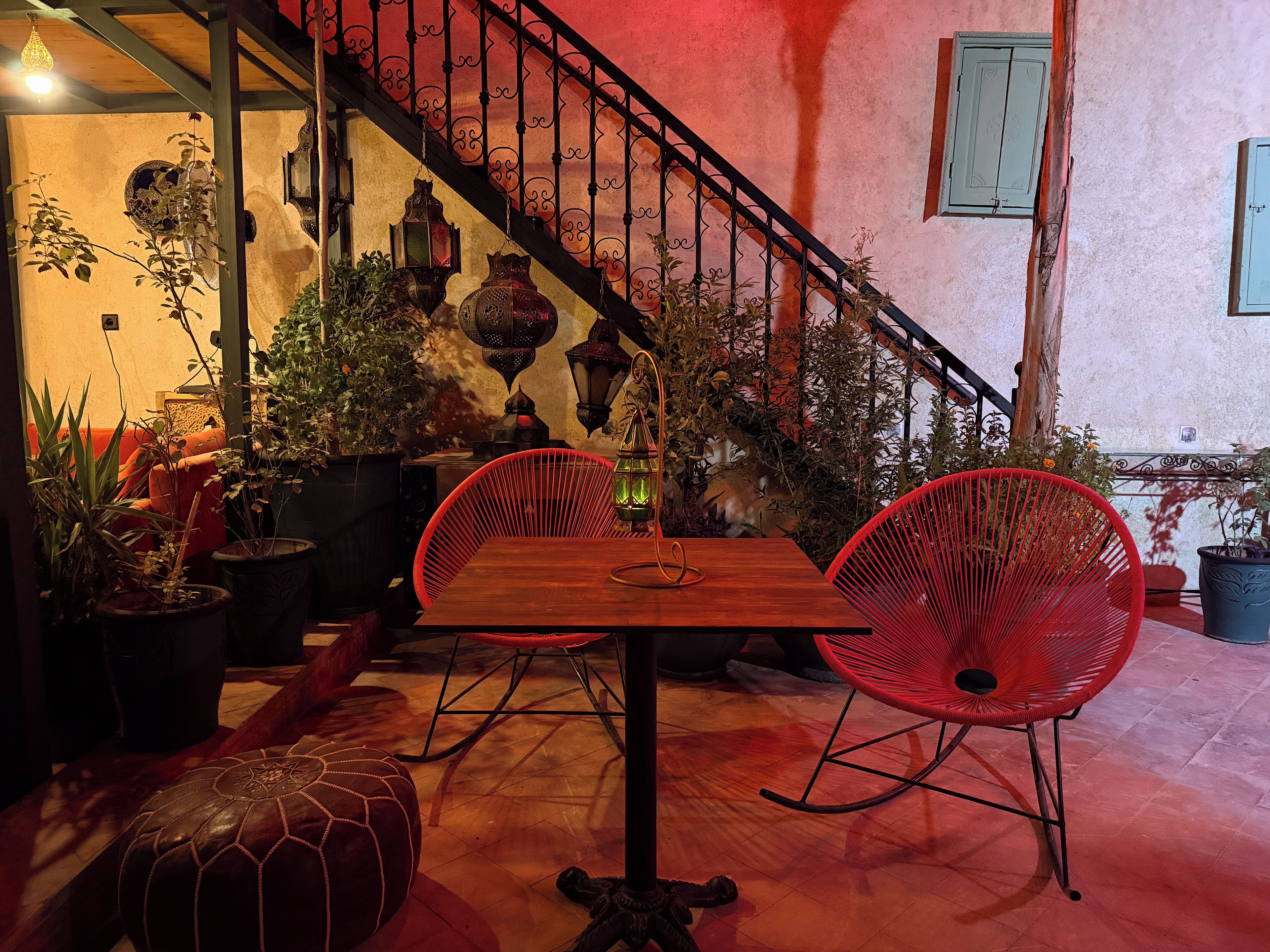Un espacio interior acogedor con una mesa de madera, sillas rojas, plantas en macetas y una escalera con barandilla de hierro forjado.
