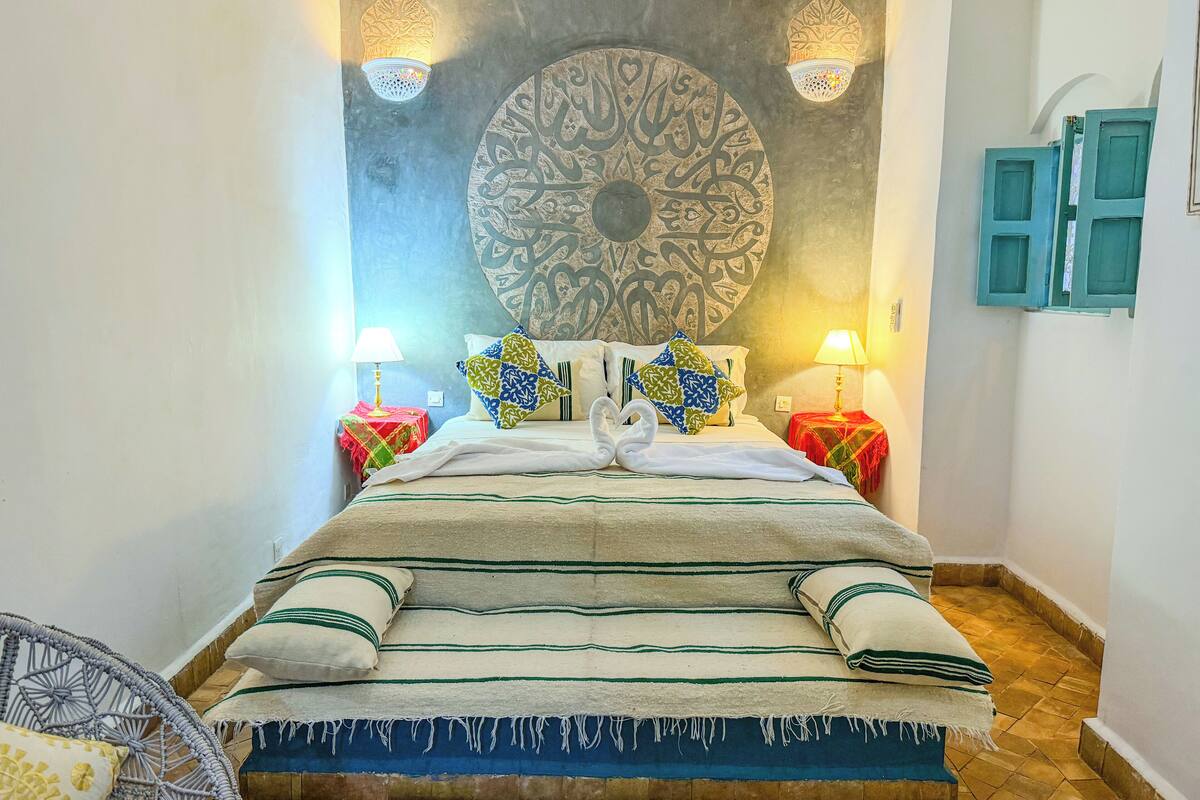 Un letto ordinato con un grande pannello decorativo a parete, due lampade fissate alla parete e un tavolino con lampada.