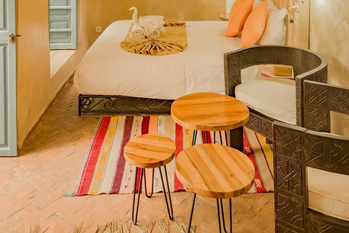Camera d'albergo con un letto, due sgabelli di legno, un tavolino, un termosifone e una lampada a parete.