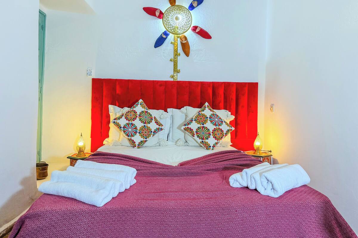 Un letto rifatto con cura, con una testiera rossa, due cuscini decorativi e una coperta a motivi.