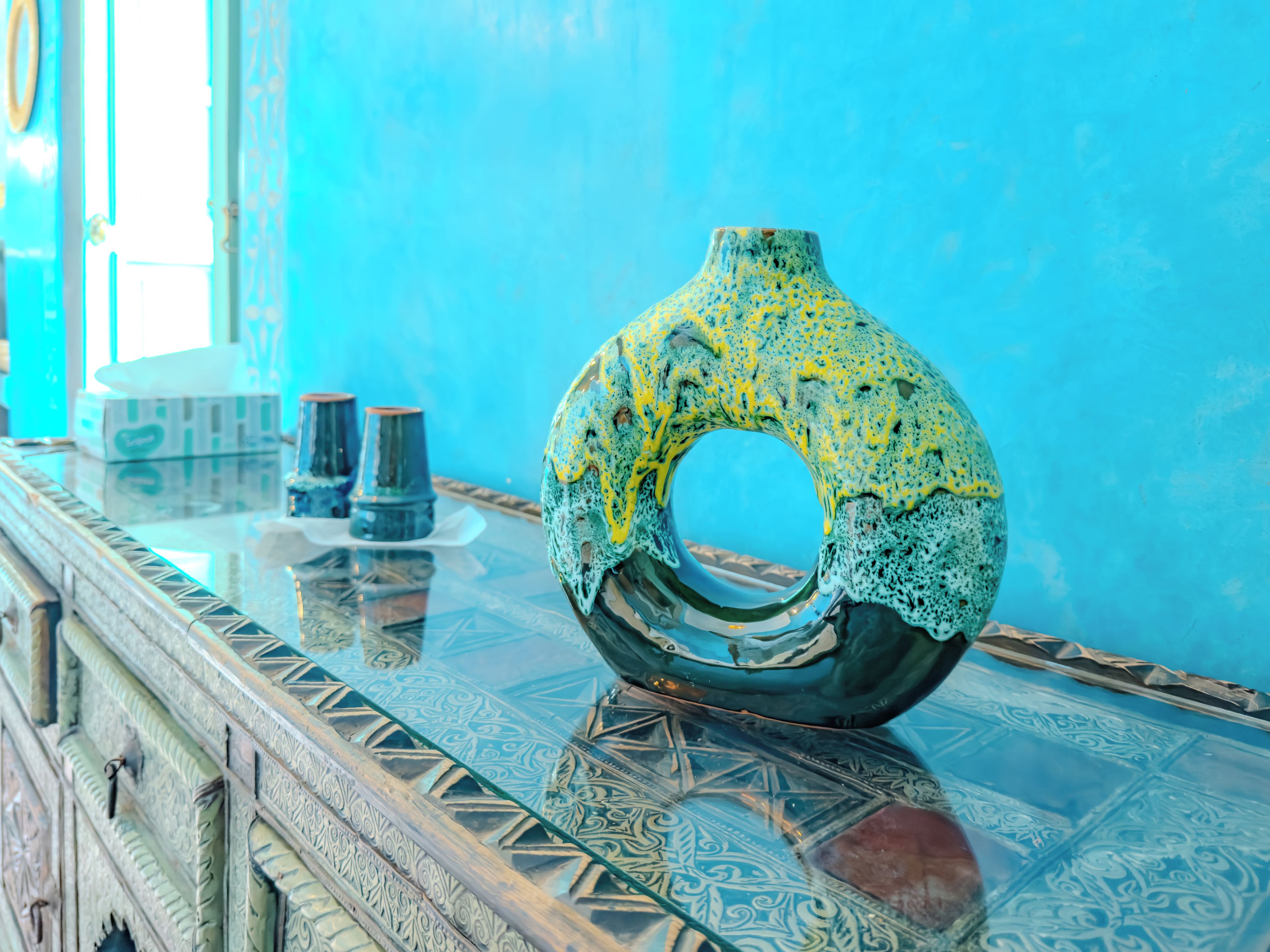 A decorative glass vase on a reflective surface with intricate patterns, set against a blue wall.