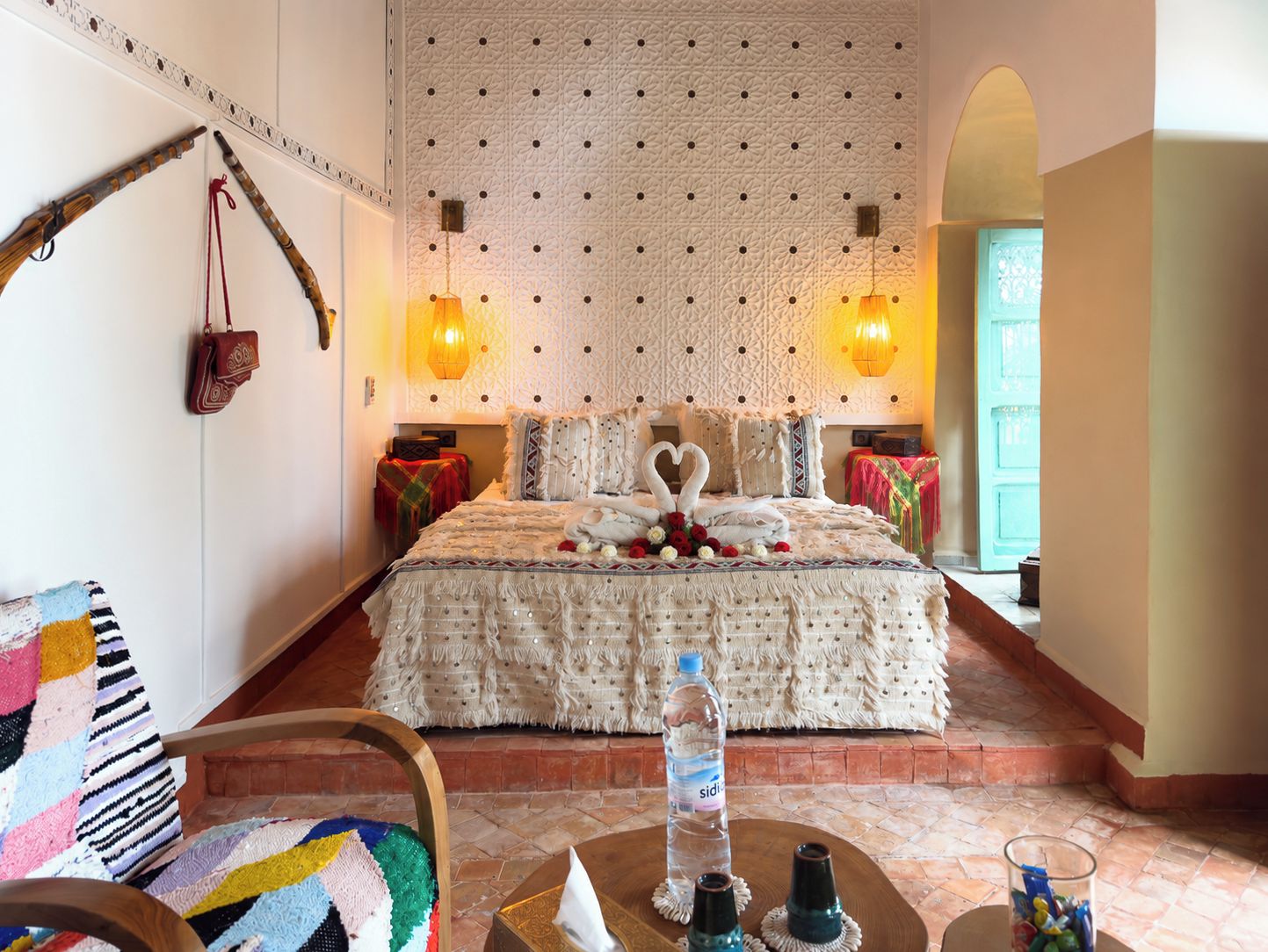 A hotel room with a bed, a chair, a table with a bottle and glasses, and a wall with a patterned design.