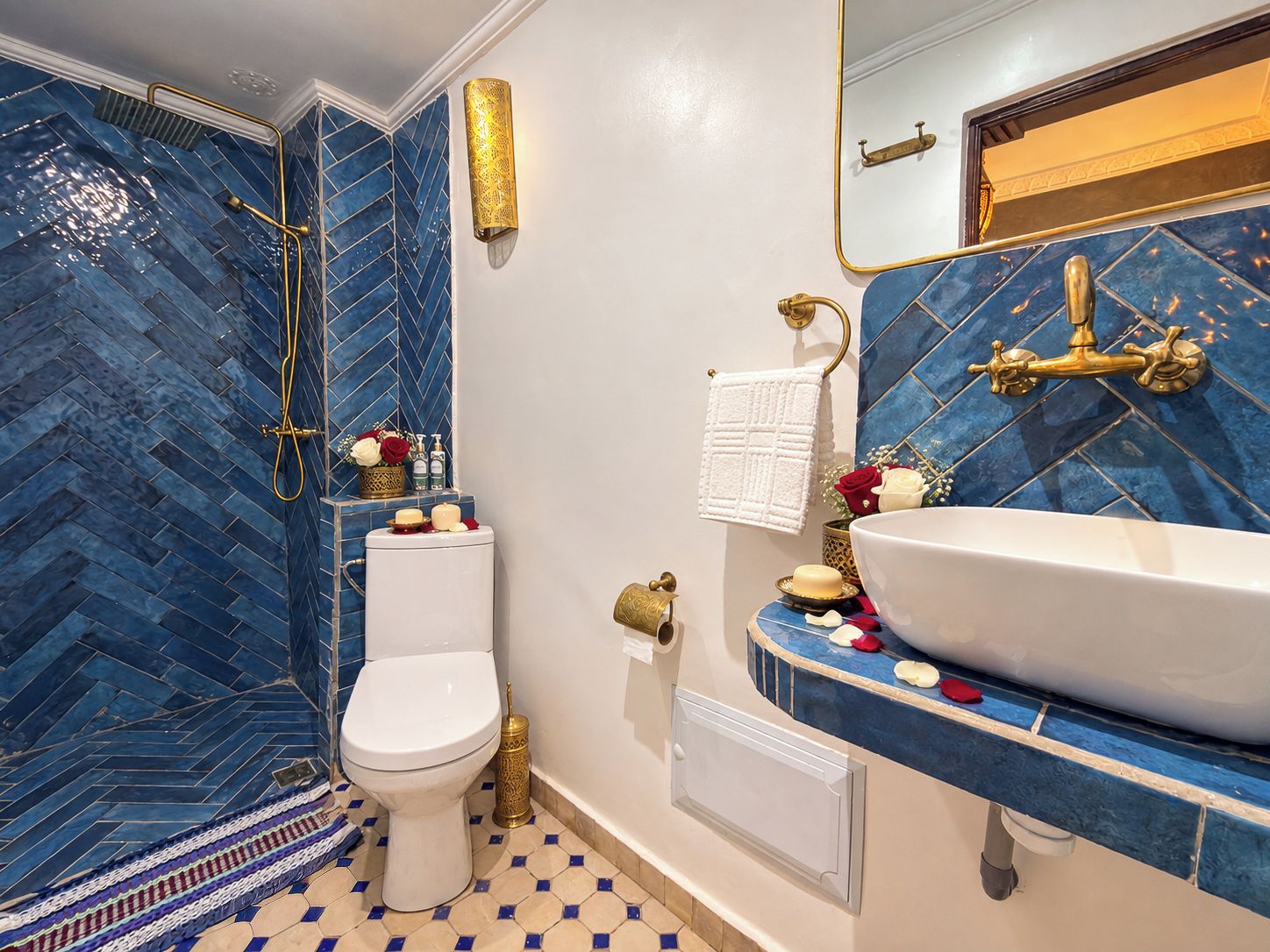 A bathroom with a blue herringbone tile backsplash, a white toilet, a gold showerhead, and a white sink with a gold faucet.