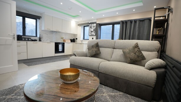 A modern living room with a sofa, a coffee table, and a kitchen area with cabinets and a refrigerator. - Charming London House - Sleeps 4 & Outdoor Space (London)