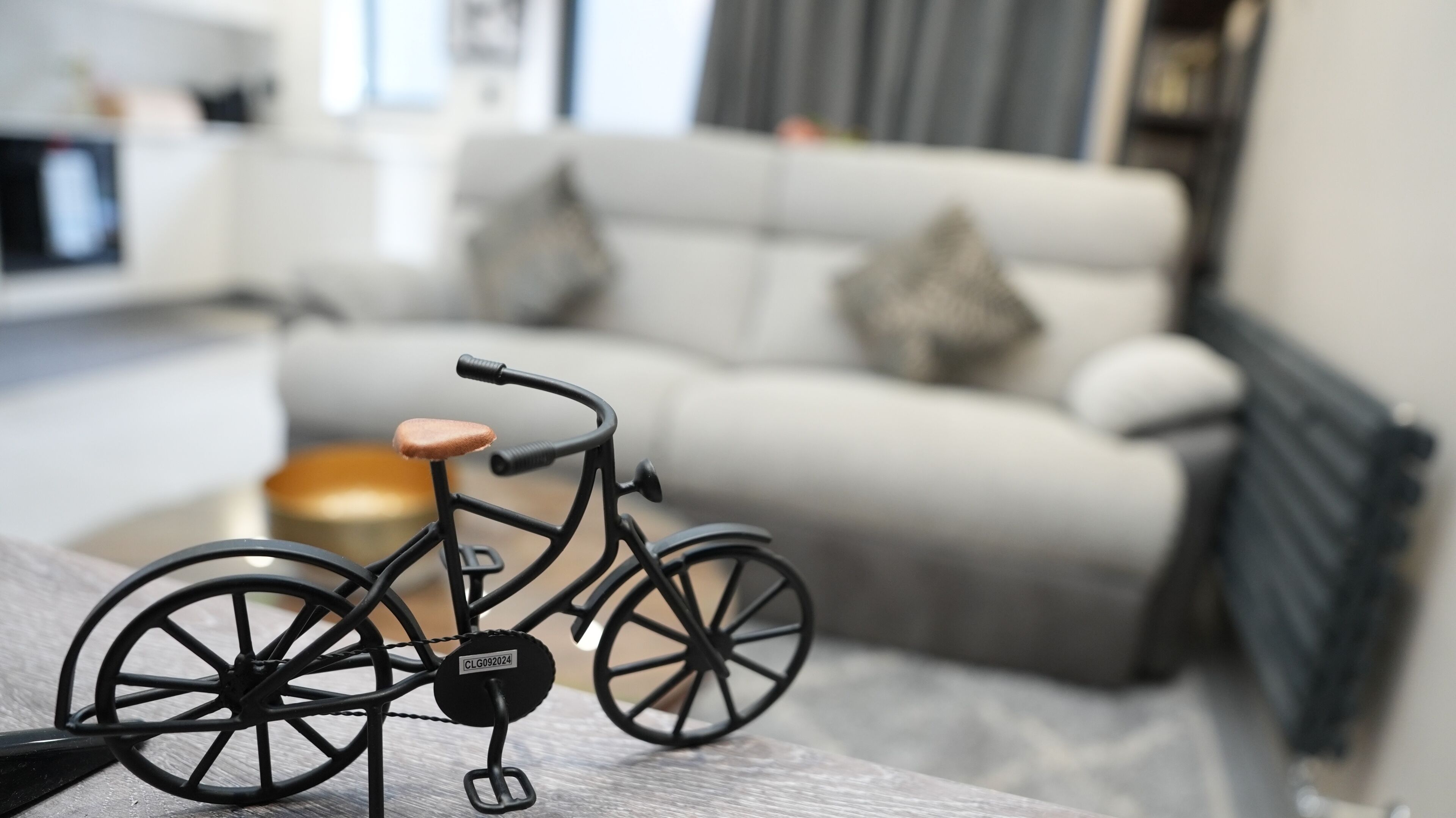 A model bicycle with a tag on the front wheel, placed on a table in a modern living room with a sofa and a radiator in the background.