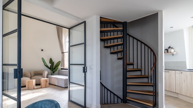 Un intérieur moderne avec un escalier en colimaçon, un coin salon agrémenté d’une plante et un espace cuisine.